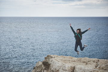 Wall Mural - Freedom. Full length portrait of happy girl is jumping on rock mountain with excitement. She is stretching hands up and laughing. Wonderful seascape on background 