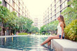 Summer in the city. Happy young woman sitting near condominium swimming pool.