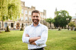 © Drobot Dean - Portrait of adult smiling man 40s in white shirt, looking at camera during walk in green park while standing with arms folded over old building background