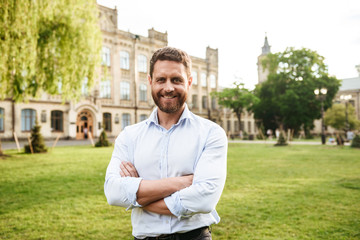 Portrait of adult smiling man 40s in white shirt, looking at camera during walk in green park while standing with arms folded over old building background