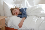 Fototapeta Młodzieżowe - Petit garçon dort paisiblement dans son lit, visage détendu, cheveux longs en bataille sur l'oreiller blanc, bouche légèrement ouverte 