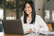 Image of joyful asian female worker 20s wearing white shirt smiling while sitting at table in office, and working on laptop