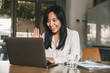 © Drobot Dean - Image of happy asian woman 20s wearing white shirt smiling and waving hand at laptop, while speaking or chatting on video call in office