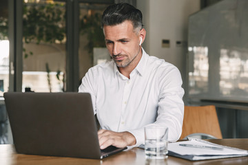 Fototapete - Photo of european businessman 30s wearing white shirt and wireless earphones sitting at table in office, and working at laptop