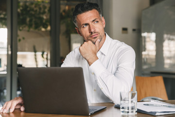 Fototapete - Photo of european man wearing white shirt and earbud sitting at table in office with brooding look aside, while working on laptop