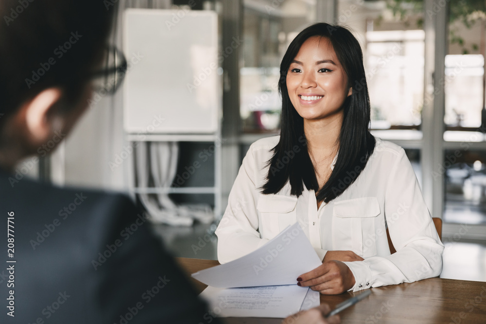 Portrait of joyful asian woman smiling and holding resume, while sitting in front of businesswoman during corporate meeting or job interview - business, career and placement concept - obrazy, fototapety, plakaty 