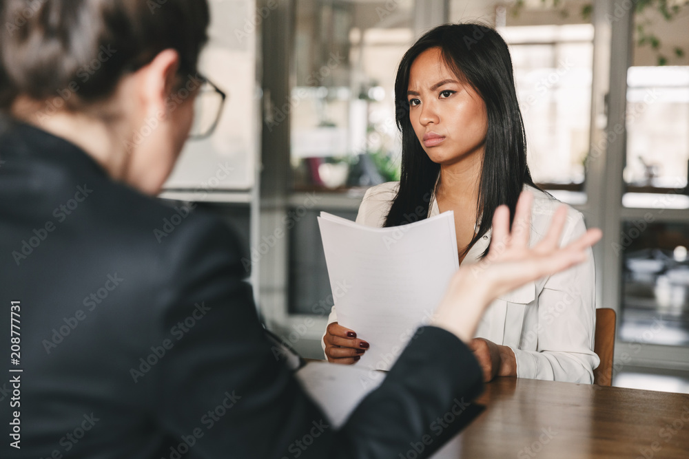 Image of serious asian woman looking and talking to businesswoman, while sitting at table in office during job interview - business, career and recruitment concept