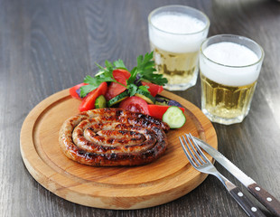 The sausage in the form of a spiral is cooked on the grill. Next to a salad of fresh vegetables. In the frame cutlery and two glasses of beer. Dark background. Close-up.