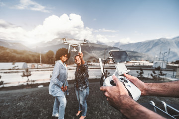 Two cheerful girls of different races are posing for broadcast from the drone which is filming them in a defocused background with mountains; male hands with the remote controller in the foreground