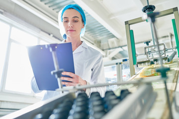 Wall Mural - Low angle portrait of  young female worker wearing lab coat standing by  power units and writing on clipboard  in clean production workshop, copy space