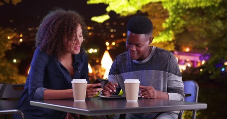 Wall Mural - Attractive black male and female using tablet computer outdoors at night, Friends meeting for coffee using modern technology and talking, 4k