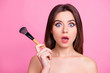 Close up portrait of shocked beautiful attractive pop-eyed pretty surprised scared make up artist holding a brush for blusher and powder in a hand, isolated on pink background
