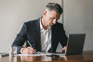 Fototapete - Image of concentrated confident businessman 30s wearing white shirt and black suit working at laptop in office, while writing down notes