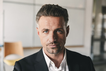 Fototapete - Portrait closeup of serious concentrated businessman wearing white shirt and black suit looking at camera, while working in office room