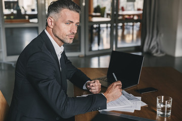 Fototapete - Image of concentrated confident businessman 30s wearing white shirt and black suit sitting at table in office, during work with documents and laptop