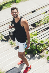 Wall Mural - high angle view of happy sportsman holding bottle of water and bag for sport equipment on stairs at sport playground