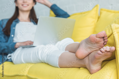 Fotomural cropped shot of smiling barefoot girl using laptop on sofa at home