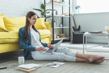 concentrated barefoot girl using laptop while sitting on carpet and studying at home