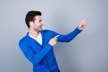 Side view portrait of cheerful positive guy in blue cardigan pointing with two forefingers, isolated on grey background