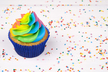 Cupcake with rainbow colorful cream in blue cup on white wooden table decorated with colorful sprinkles.