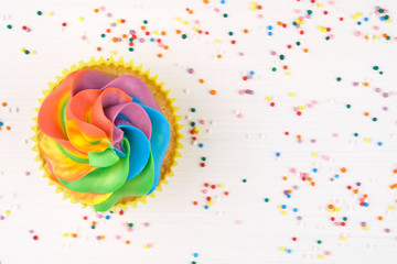 Wall Mural - Cupcake with rainbow colorful cream in yellow cup on white wooden table decorated with colorful sprinkles. Top view.