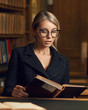 Beautiful blonde woman wearing elegant black tweed jacket and glasses sitting at desk beside bookshelf and reading book. Young gorgeous female student studying at library. Smart is new sexy concept.