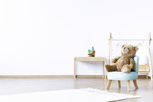 Teddy Bear On Blue Chair In White Boy's Room Interior With Wooden Table And Copy Space. Real Photo