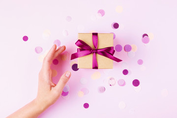 Wall Mural - Woman hand and kraft gift box with pink bow on pink background decorated with confetti.. Top view, holiday present concept.