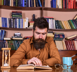 Leinwandbilder - Man on concentrated face reading book, studying, bookshelves on background. Self education concept. Teacher or student with beard studying in library. Man sits at table with mug and hourglass.