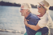 Happy Senior Couple Have Fun And Enjoy Outdoor Leisure Activity At The Beach. The Man Carry The Woman On His Back To Enjoy Together A Retired Lifestyle At The Beach