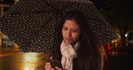 Wall Mural - Millennial woman smiling and reading text message on smartphone underneath umbrella on the street at night, Female in urban area during rainstorm in the evening using cell phone, 4k