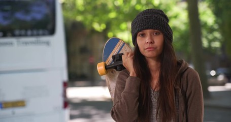 Wall Mural - Millennial girl holding skateboard over her shoulders looking at camera on the street during the day, Medium shot portrait of hipster woman with attitude standing by public bus, 4k