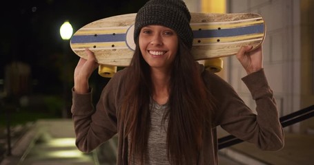 Wall Mural - Millennial girl with skateboard over her shoulders looking at camera at night, Medium shot portrait of hipster woman with attitude holding shortboard on sidewalk, 4k