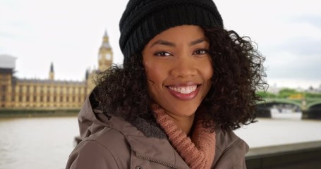 Wall Mural - Close up of happy African American woman smiling at camera in front of Big Ben wearing jacket and beanie, Black Millennial tourist on vacation in London standing by the Thames during winter, 4k