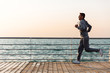 Side view of young running sportsman, during workout on quay, near the ocean. Full length.