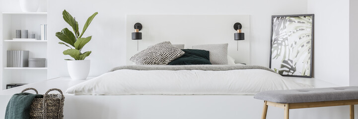 Fresh green plant standing on platform by the mattress with white bedding and patterned pillows, two lamps on bedhead and simple poster in the corner