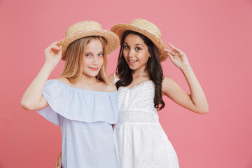 Portrait of brunette and blonde summer girls 8-10 wearing dresses posing at camera touching straw hats with smile, isolated over pink background