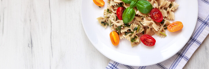 Panoramic view from above on wooden mockup table, white plate with farfalle aglio olio pasta, cherry tomatoes and fresh basil