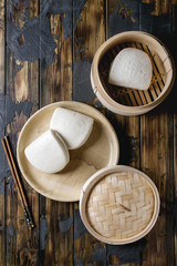 Wall Mural - Empty gua bao steamed buns in ceramic plate and opened bamboo steamer over dark wooden plank background. Flat lay, space. Asian fast food.