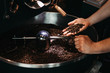 Hands of a men holding a fresh roasted bean above a metal drum full of coffee beans