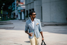 A Young And Handsome Indian Asian Man Walks Down A Street In Asia While Drinking A Coffee And Holding His Gym Bag. He Is Dressed Smartly And Fashionably In A White Shirt, Blue Jacket And Khakis. 