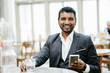 A young Indian Asian businessman laughs as he reads a funny story on his social media feed. He is seated in a cafe, taking a break from a tiring day at work.
