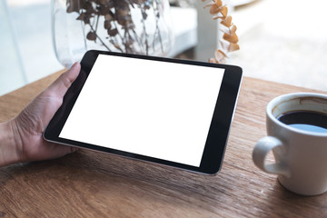 Mockup image of hand holding black tablet pc with white blank screen and coffee cup on table background