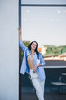 © Денис Кипкаев - A beautiful woman in a shirt and pants is standing by the mirror window of the balcony of the building.