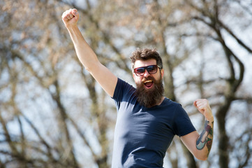 Fashion and style concept. Man with beard and mustache on cheerful face, nature background, defocused. Bearded man wears stylish sunglasses. Hipster with beard looks stylish and happy on sunny day