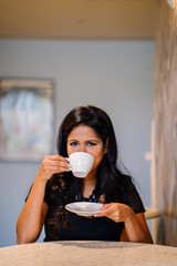 An image of a confident and successful Indian Asian Desi woman sitting at a table and wearing a professional black dress for her attire.She is drinking her coffee while waiting for her workmates.