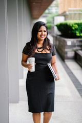 A young desi business woman grabs some coffee as she heads to her next client meeting. She looks happy as she walks along a shaded path.