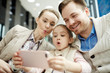 © pressmaster - Family of three grimacing in front of camera while making selfie on smartphone in lounge of airport