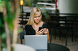 © Danon - A charming blonde lady is checking her smartphone for messages. Her laptop is on the table in front of her.