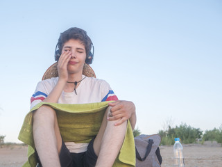 young happy boy in casual outfit with towel sitting on the beach listening to music copy space